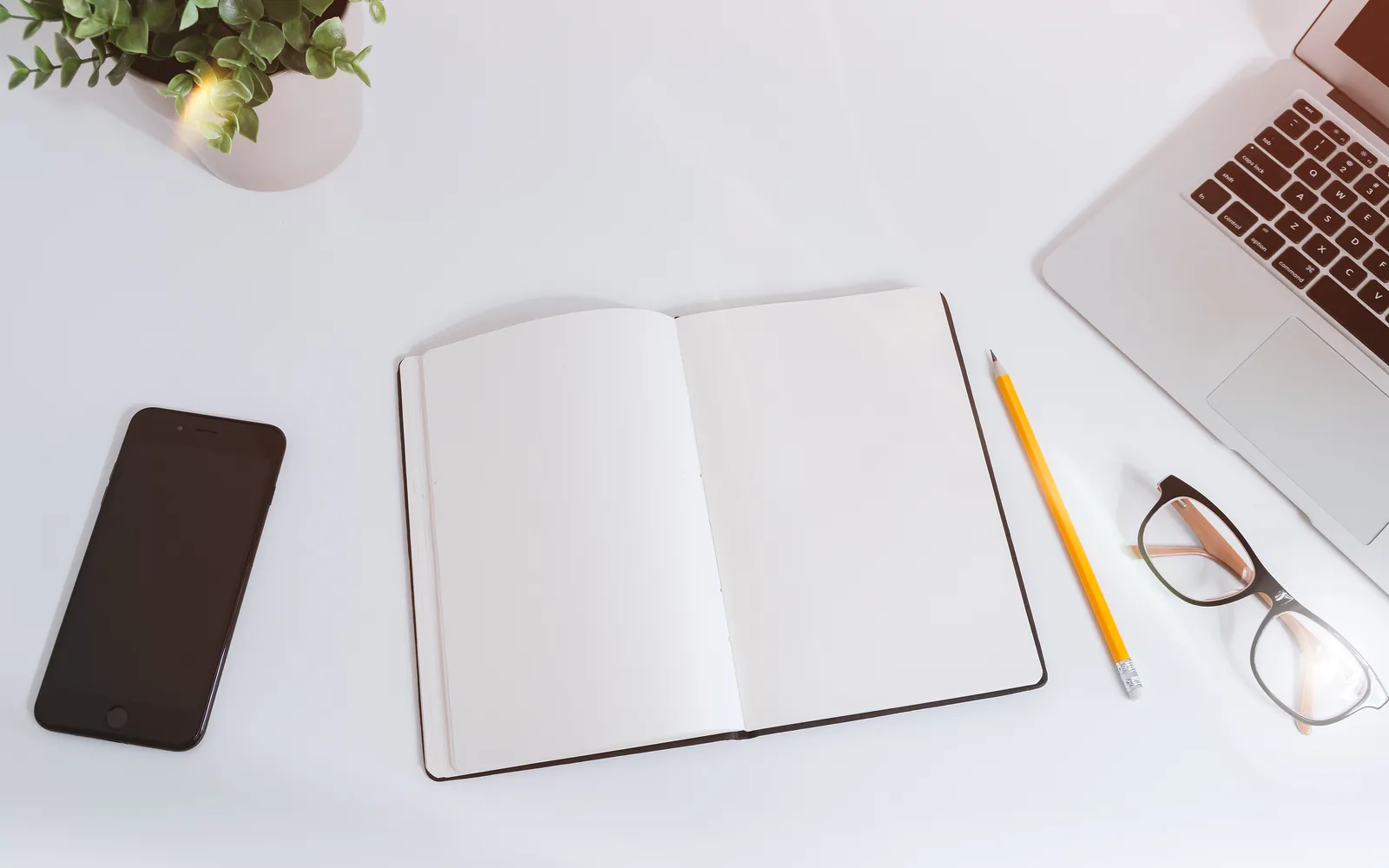 A white desk with a smartphone, an open notebook, a pencil, glasses and a laptop